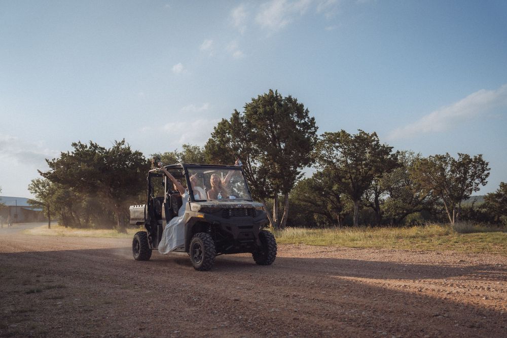 A utility vehicle drives across a dusty dirt field with trees and blue sky in the background.