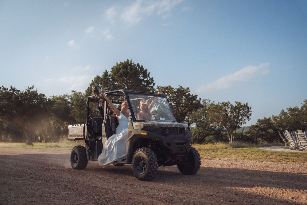 Bride in white wedding dress riding a UTV side-by-side vehicle on a dirt path at a rustic outdoor venue.