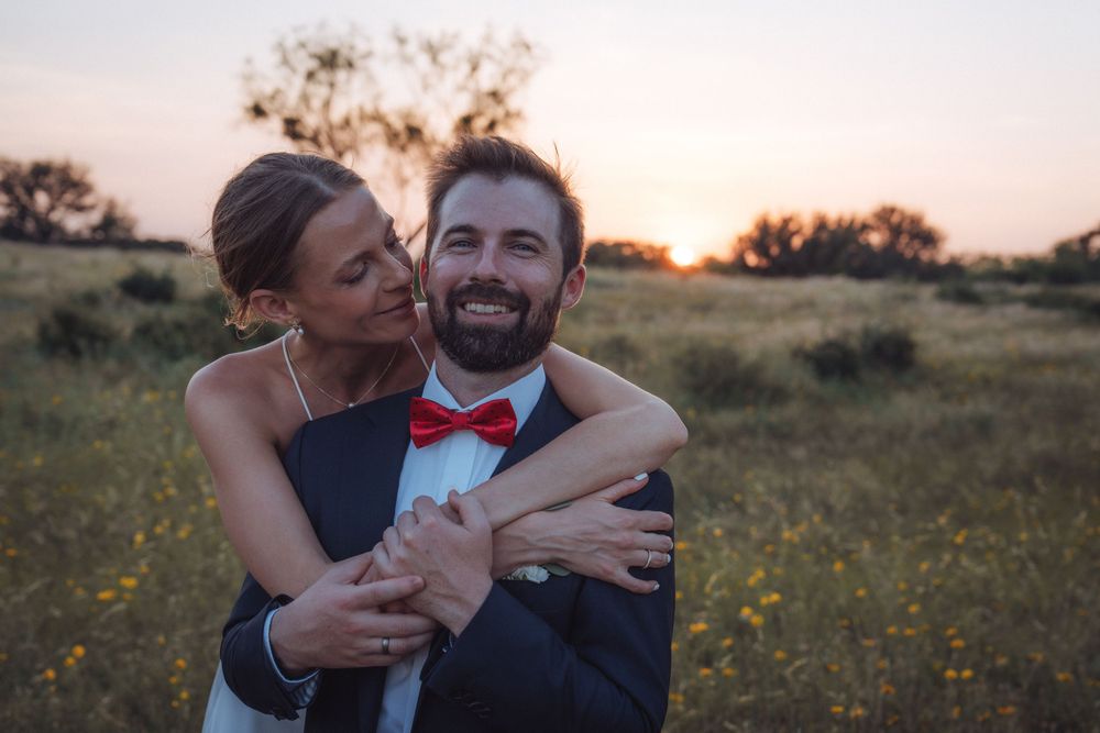 Couple embracing in a field at sunset, bride hugging groom in a navy suit with red bow tie, smiling joyfully.
