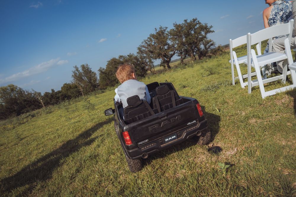 Young child riding a black toy pickup truck on green grass at an outdoor wedding venue.