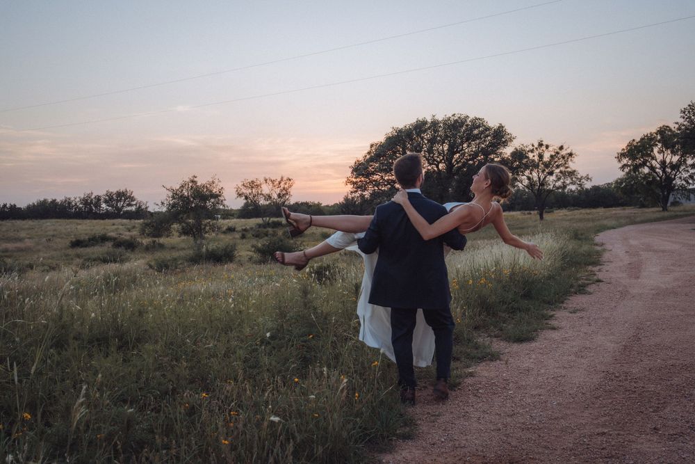 Couple dancing at sunset on a rural dirt path surrounded by lush green fields and trees in a romantic wedding photo.