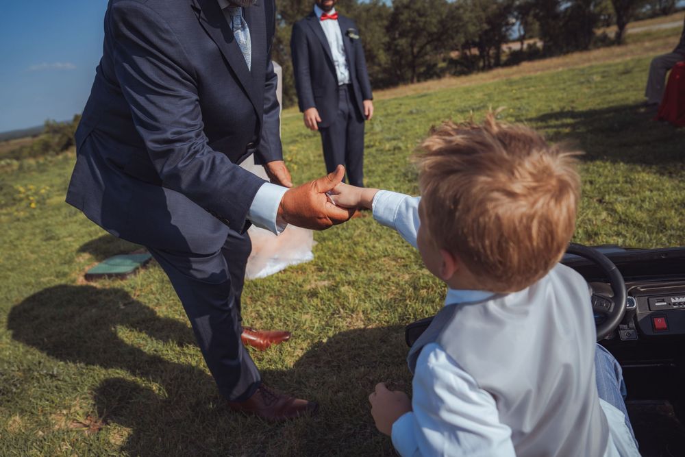 Man in navy suit shaking hands with a young boy near a toy car at an outdoor wedding.