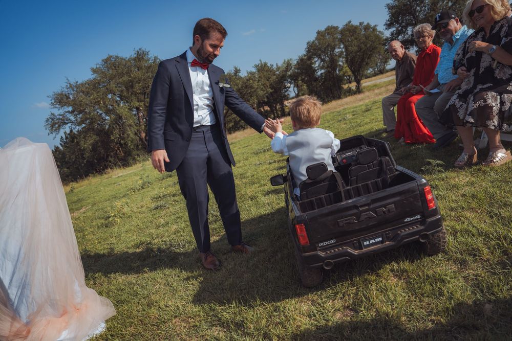 Groom in navy suit holds child's hand near toy RAM truck on green grass at outdoor wedding.