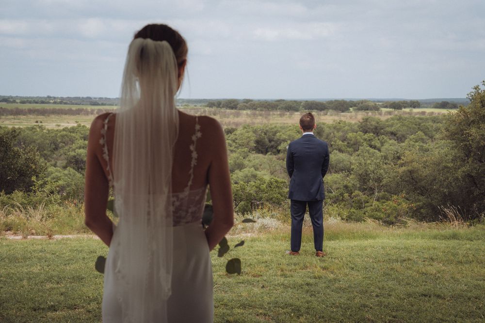 Bride in white dress approaches groom in blue suit standing at scenic overlook during outdoor wedding ceremony.