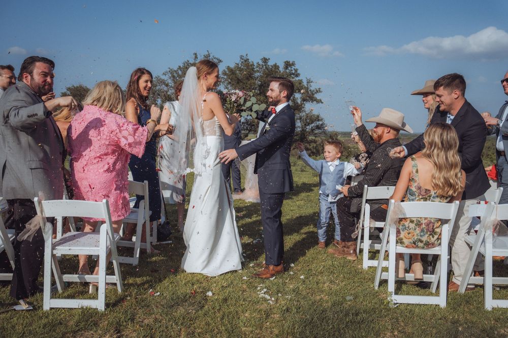 Bride and groom walk down outdoor aisle as guests celebrate at a sunny rustic wedding ceremony.