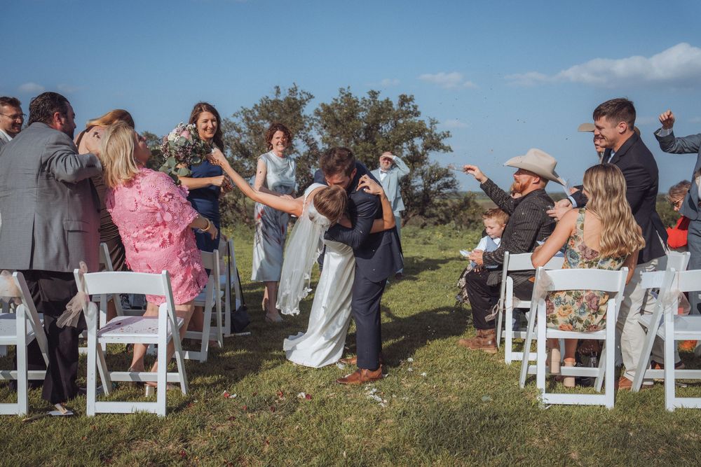 Bride and groom walk down outdoor wedding aisle surrounded by cheering guests seated in white chairs under blue sky.