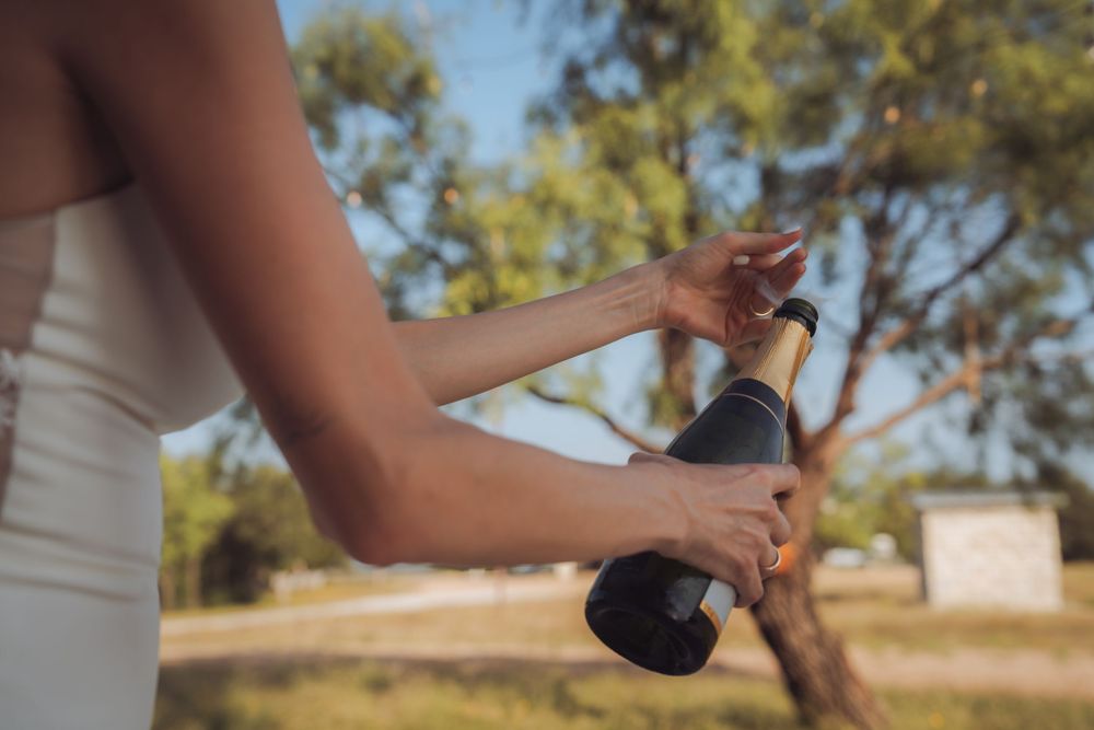Bride in white dress opening champagne bottle outdoors at wedding celebration.