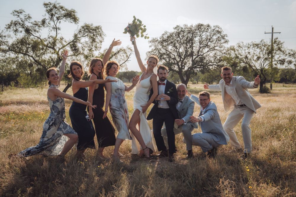 A joyful wedding party celebrates outdoors in a sunny field with oak trees in the background.