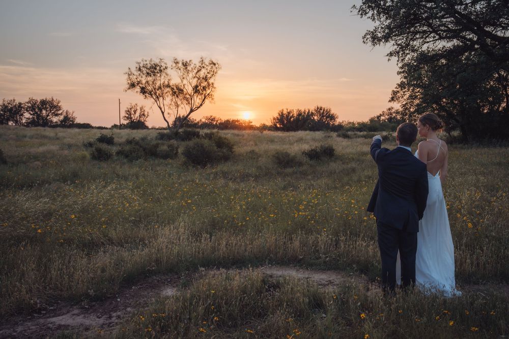 A bride and groom watch a golden sunset over a lush green field surrounded by trees.