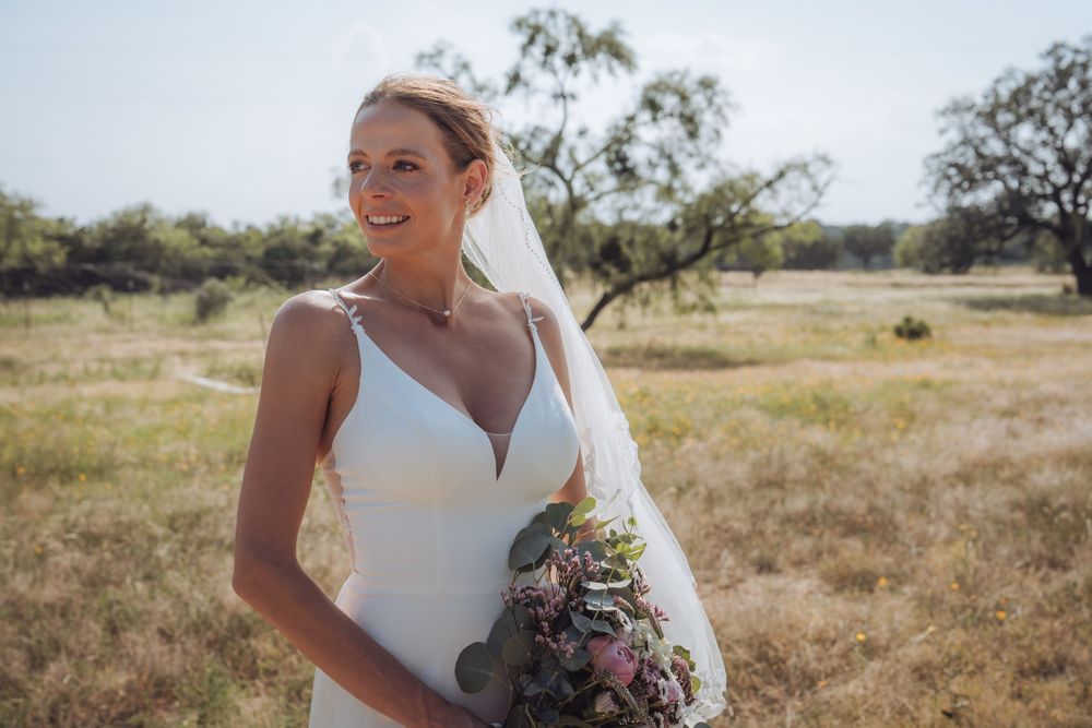Smiling bride in white gown holding floral bouquet standing in sunny outdoor field with trees in background.