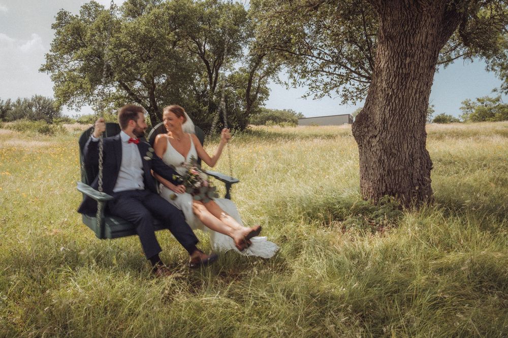 Bride and groom sitting together on a wooden swing under a large tree in a scenic green meadow on their wedding day.