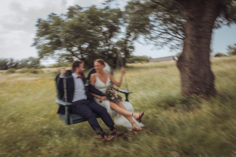 Couple in wedding attire laughing on a swing under a tree in a green meadow, captured with motion blur effect.