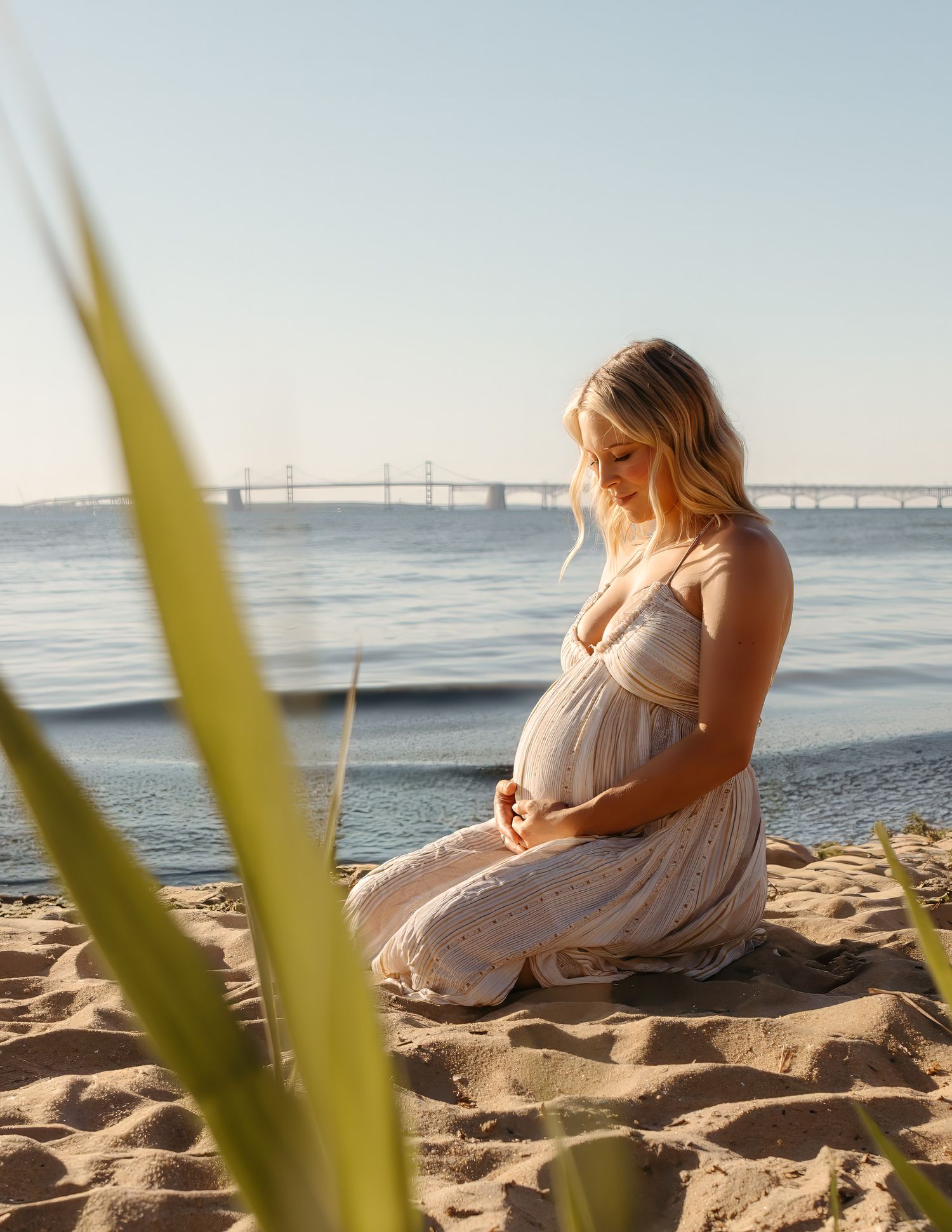 Pregnant woman in white dress sitting on sandy beach near water, cradling baby bump in golden sunlight.