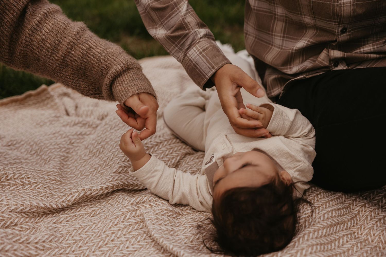 A baby lies on a blanket outdoors while a parent gently holds their hands during a family photo session in North Bend