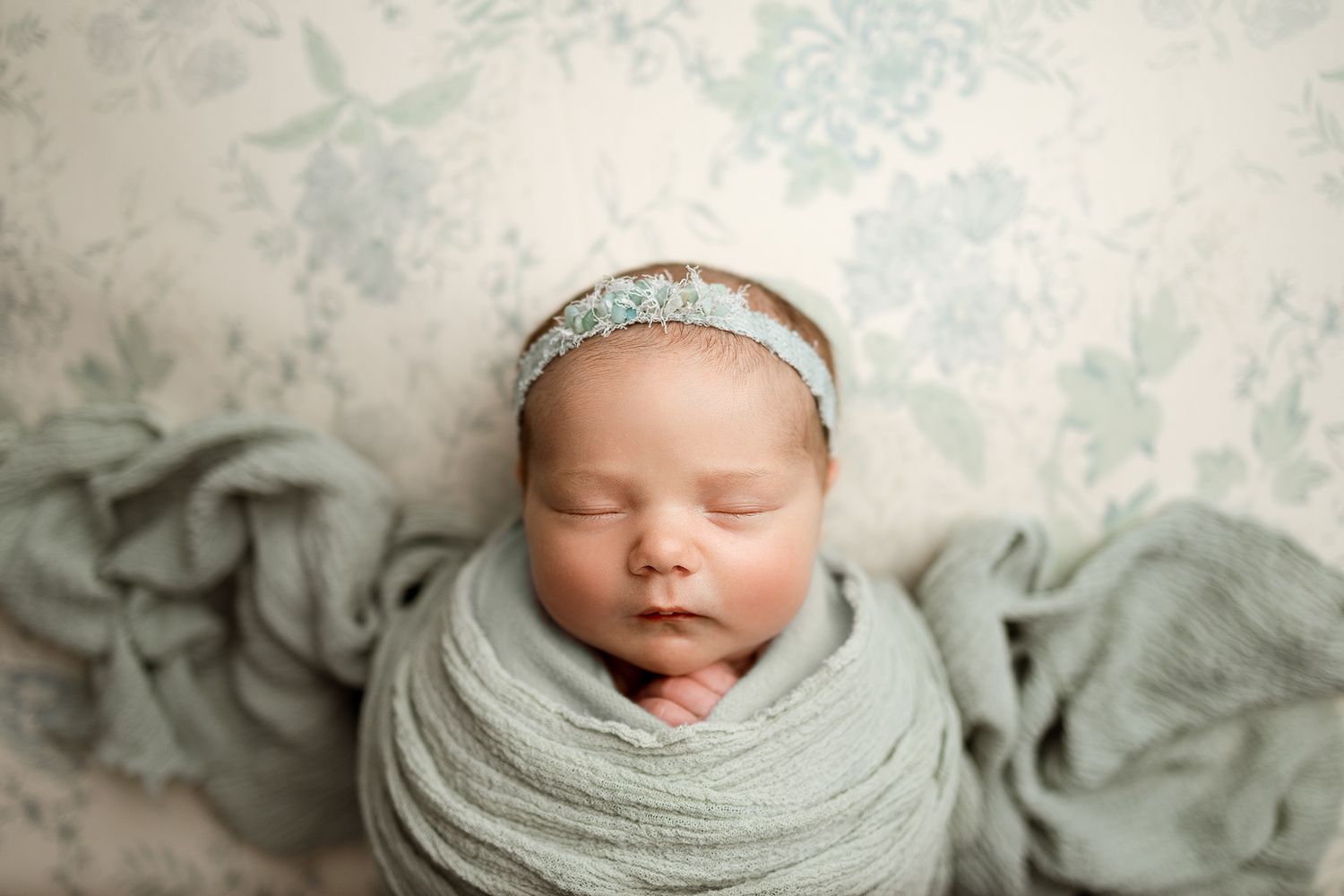 Newborn baby swaddled in sage green wrap with floral headband, sleeping peacefully on matching floral backdrop.