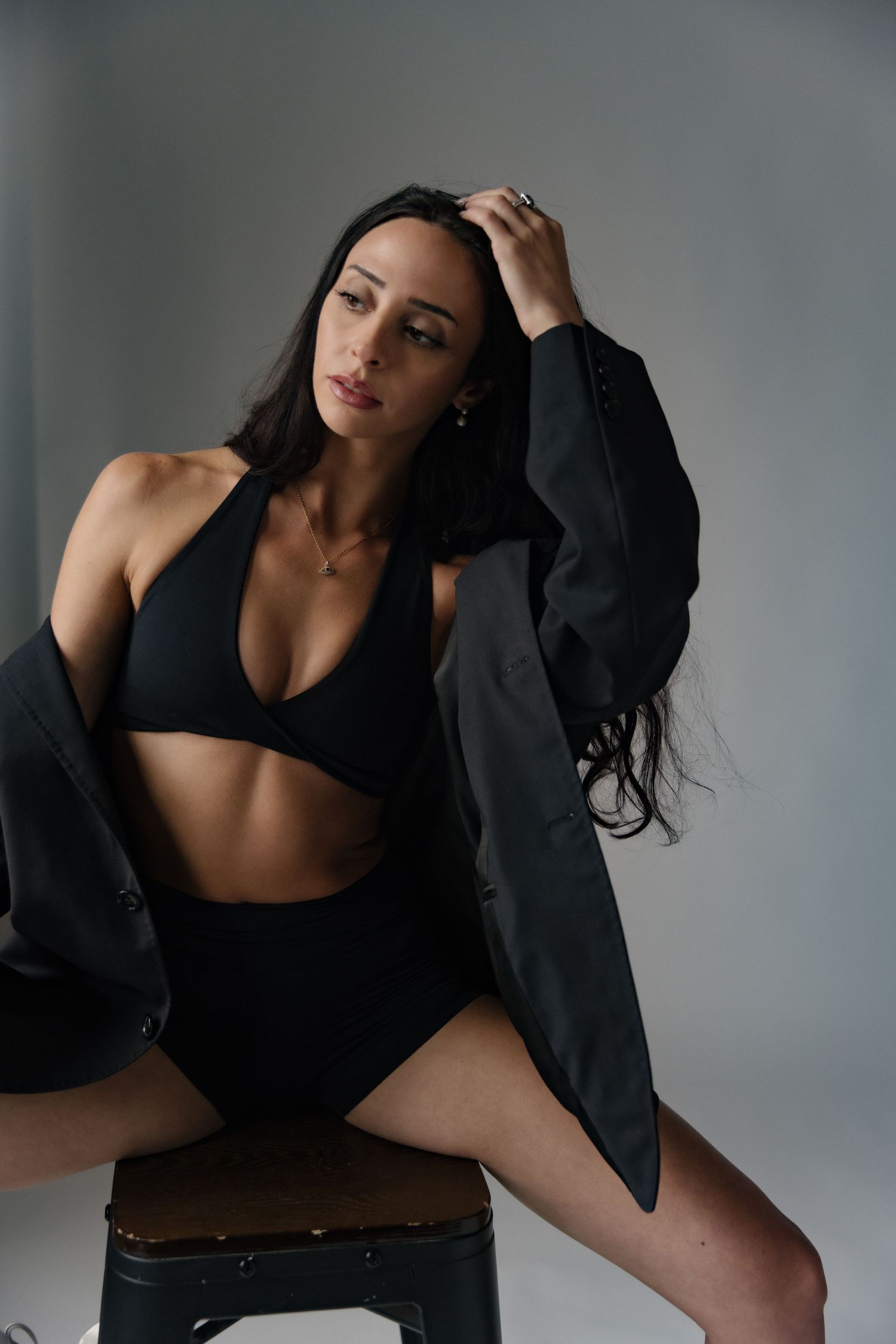 Woman in black bikini top and shorts wearing an oversized blazer, posing on a stool against a gray background.