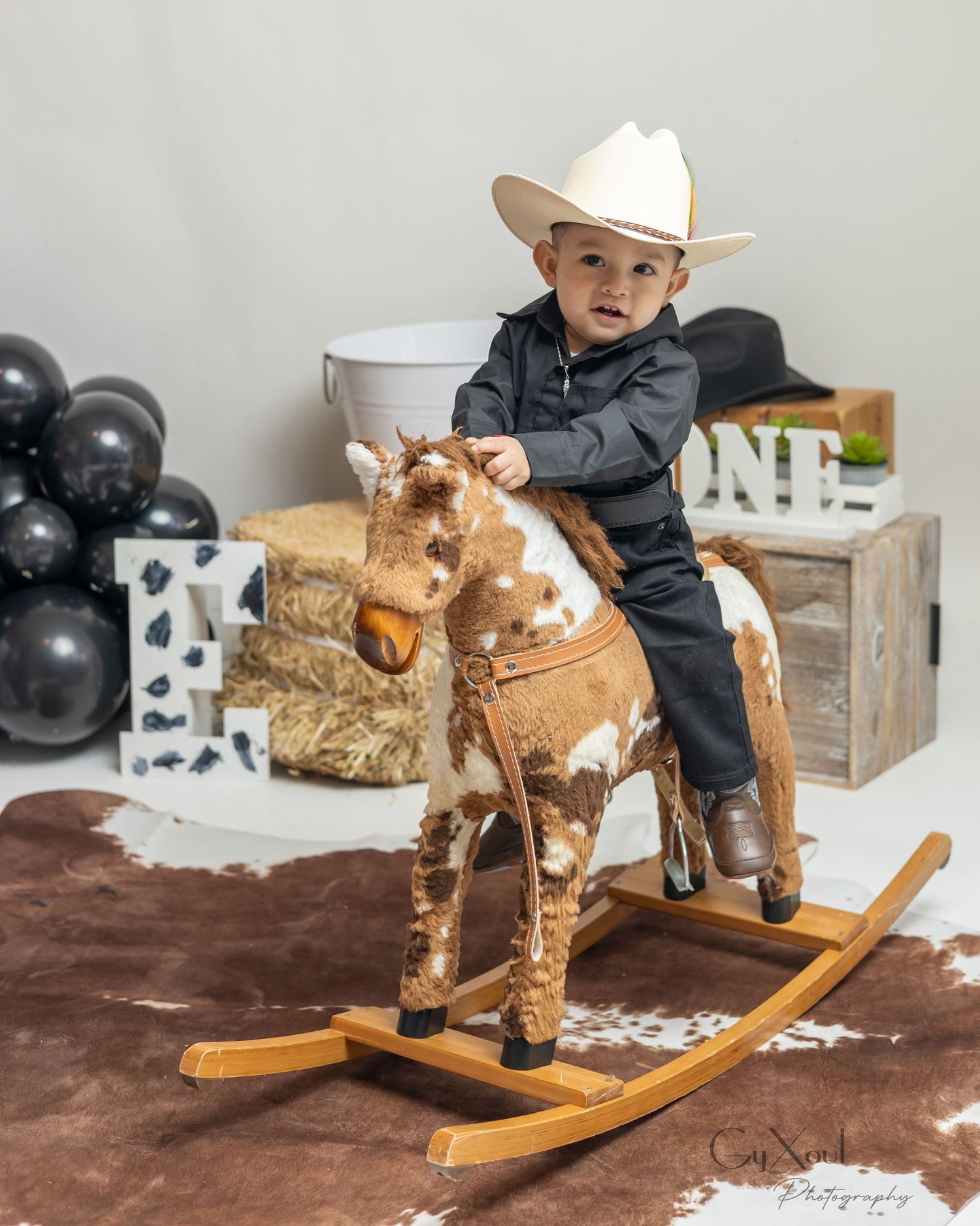 Toddler boy in black cowboy outfit and white hat riding a brown and white rocking horse at a western first birthday party.