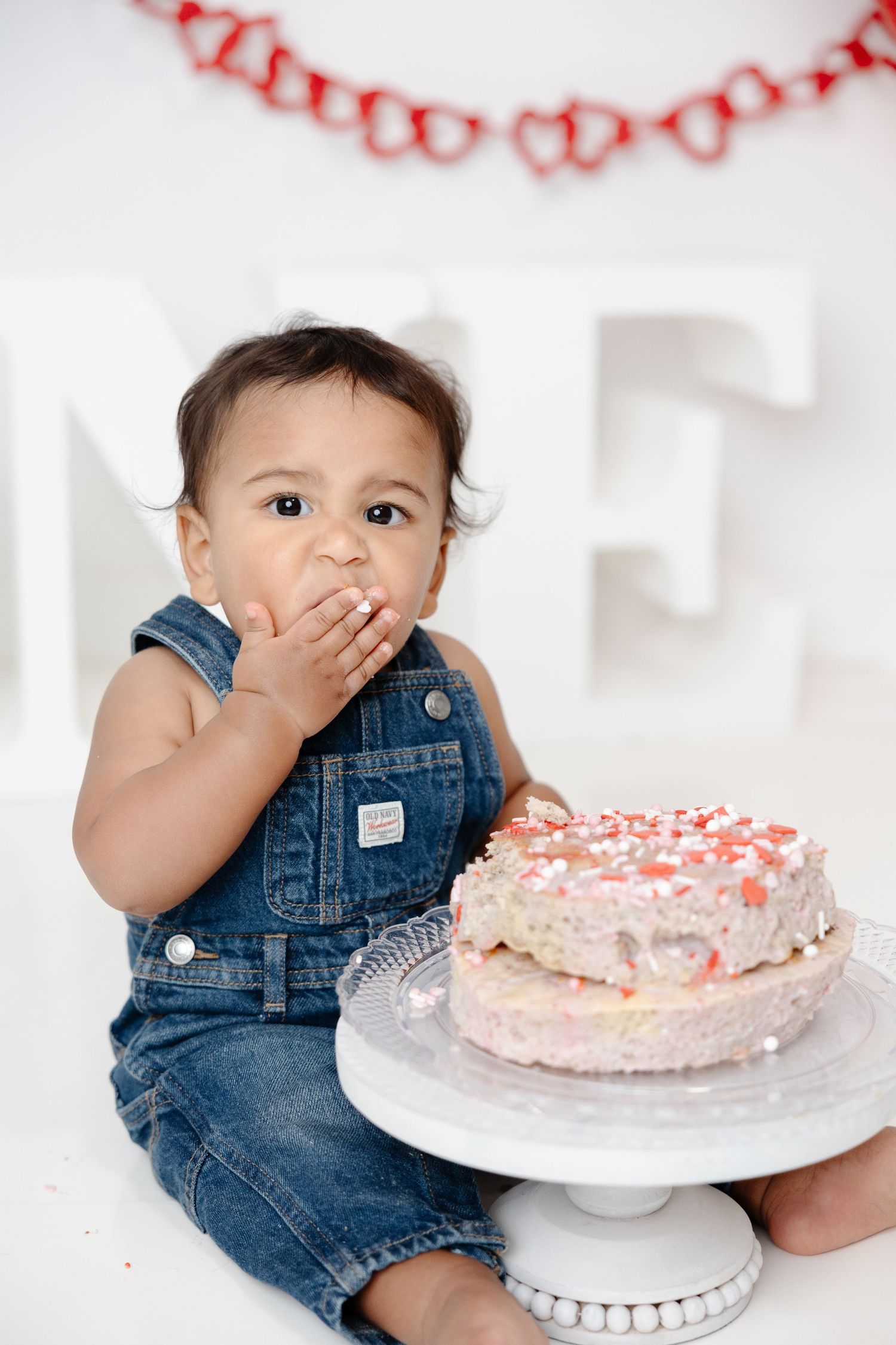 A baby in denim overalls smashes their first birthday cake on a white stand, with heart garland decor behind them.
