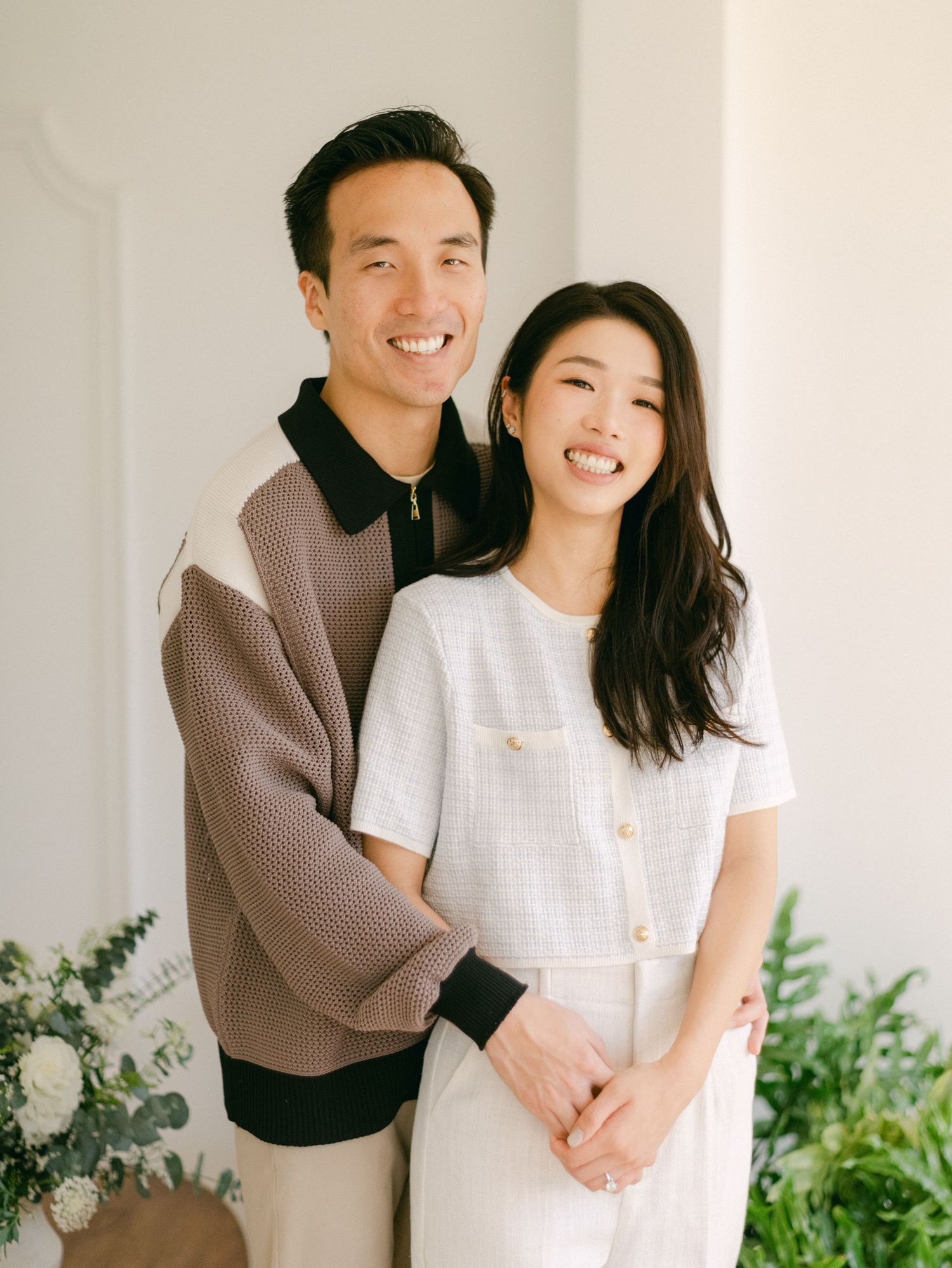 A smiling couple poses together, man in colorblock sweater embracing woman in white tweed outfit, with greenery backdrop.
