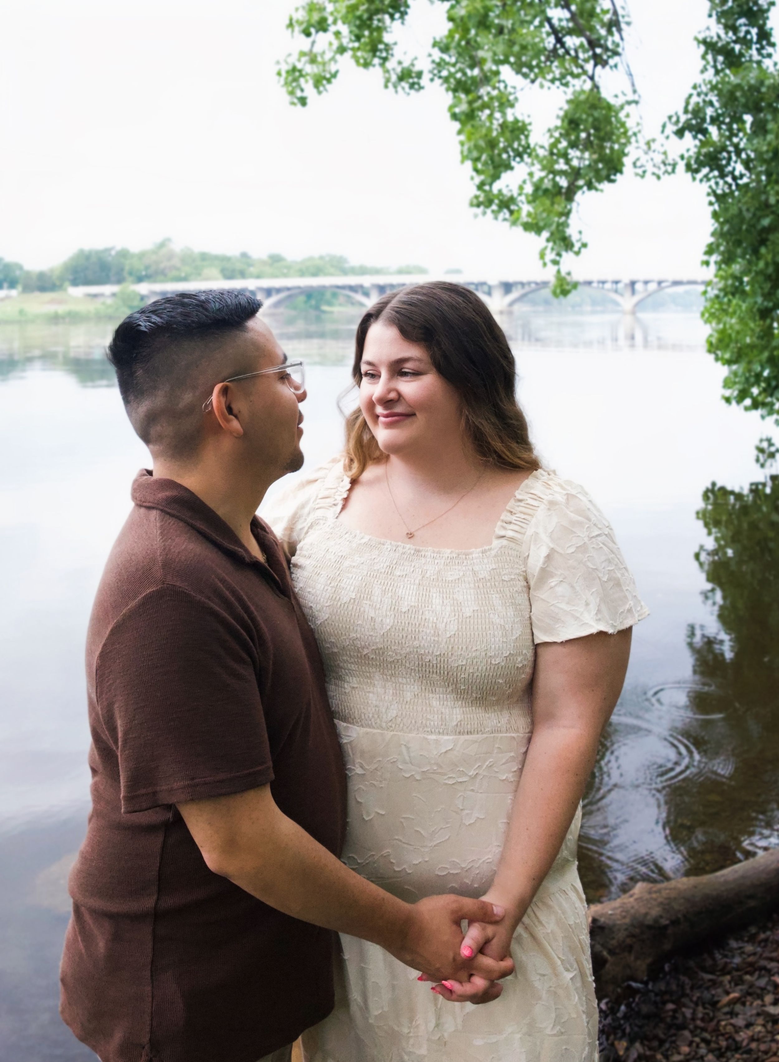 Couple holding hands and smiling at each other by a river, woman in white lace dress, man in brown polo shirt.