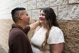 A couple smiles at each other while standing against a brick wall outdoors.