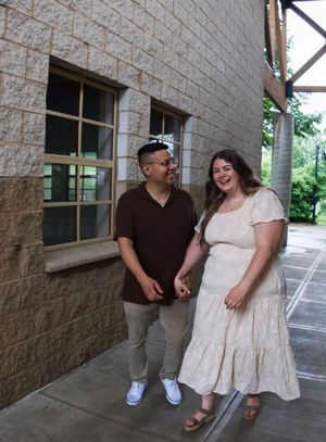 A couple smiles and holds hands outside a brick building, man in brown shirt and woman in cream dress.