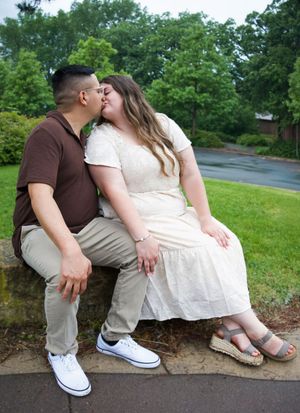 Couple sharing a kiss while sitting on a stone wall in a lush green park setting.