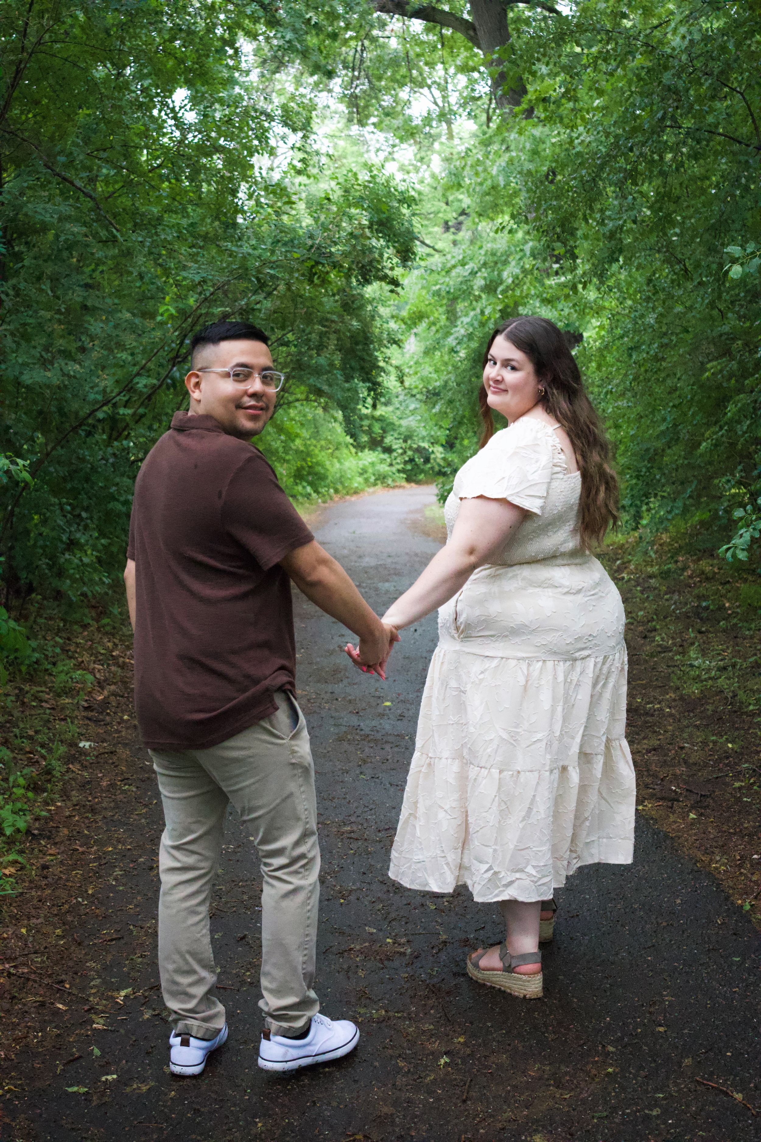 Couple holding hands on a tree-lined path, man in brown shirt and khakis, woman in cream dress, both smiling.