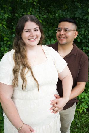 A smiling couple poses outdoors in front of lush green foliage, woman in white dress, man in brown shirt holding hands.