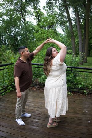 Couple dancing on a wooden deck surrounded by lush green trees, man in brown shirt twirling woman in white dress.