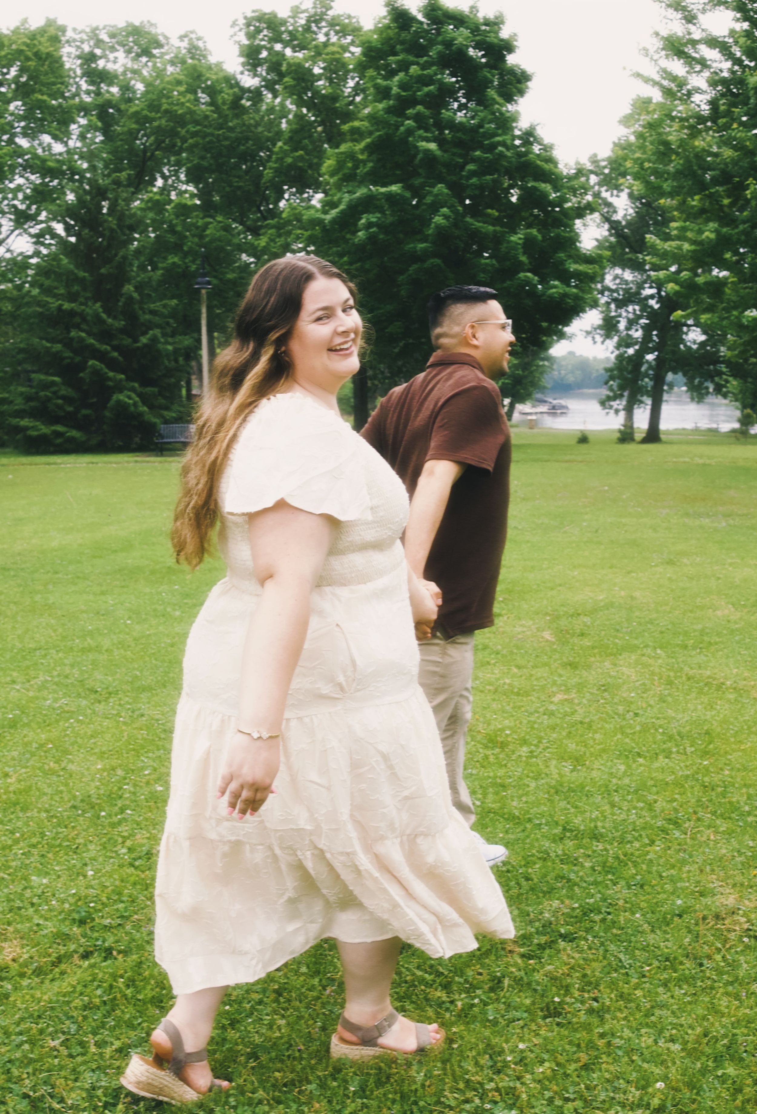 A smiling woman in a white dress walks with a man in a brown shirt across a lush green park.