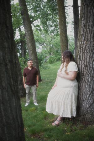 A couple poses among tall trees in a lush green park, woman in white dress leaning on tree, man in brown shirt smiling.