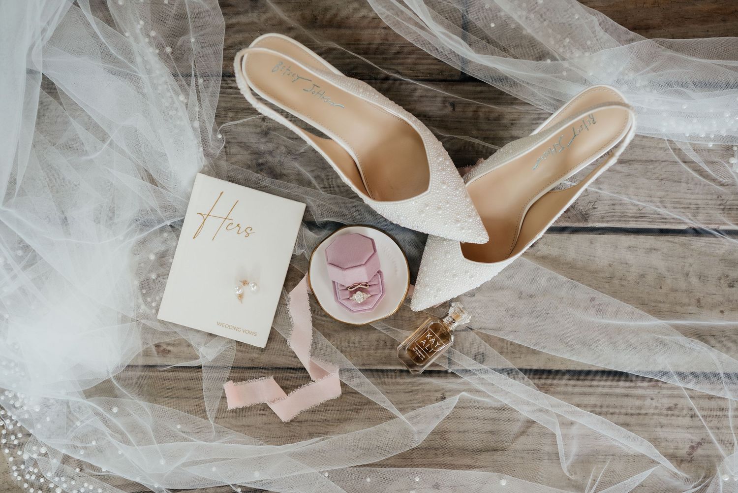 Bridal flat lay featuring white heels, veil, ring dish, perfume, and wedding stationery on wood.