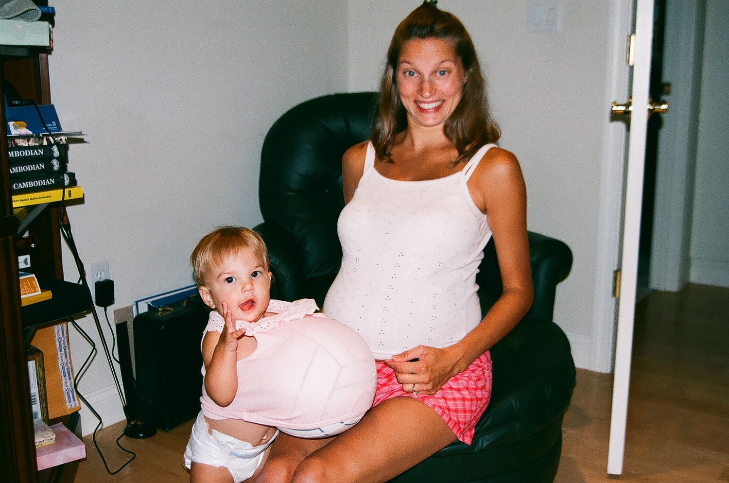 Expectant mother in white tank top sits in black chair with toddler in diaper with a ball under shirt posing as a pregnant.