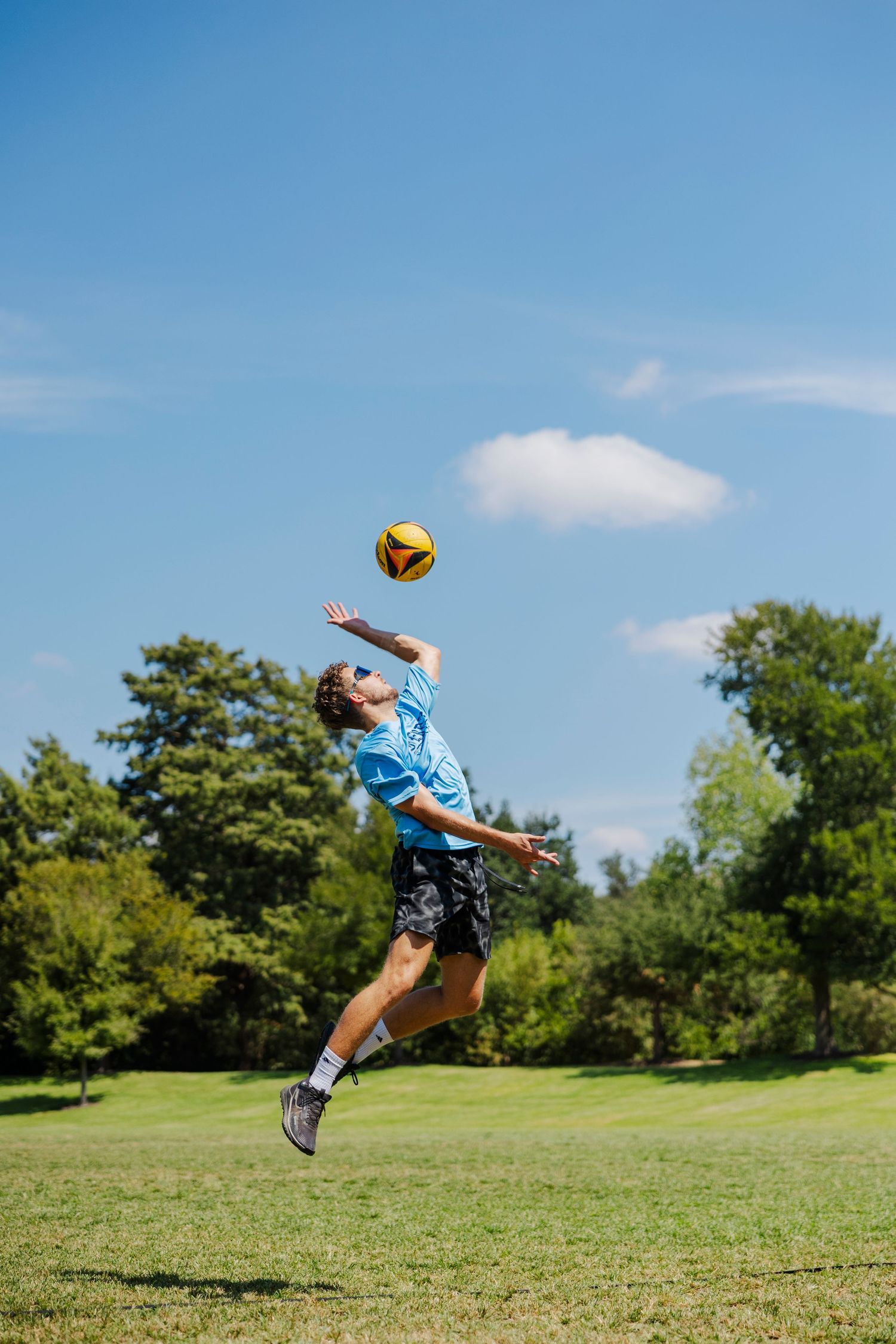 Young man jumping to hit a yellow soccer ball in a green park under a bright blue sky.