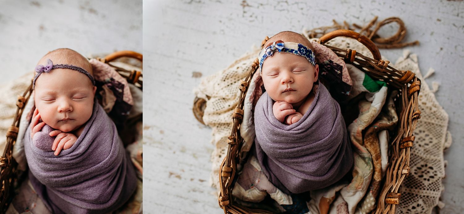 Sleeping newborn baby girl wrapped in purple swaddle with floral headband resting in a rustic wicker basket.