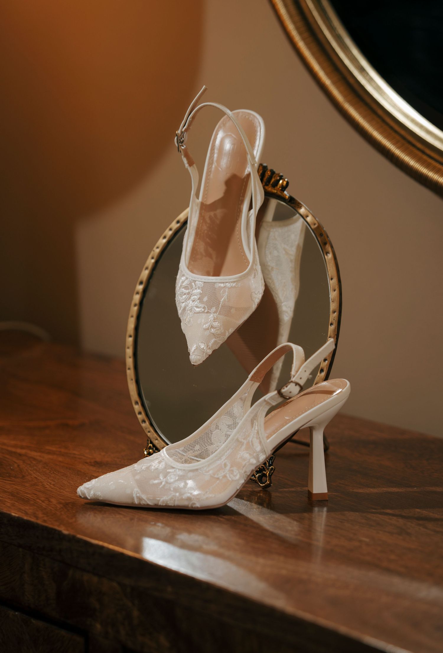 White lace slingback heels with floral embroidery displayed against a round gold mirror on a wooden surface.