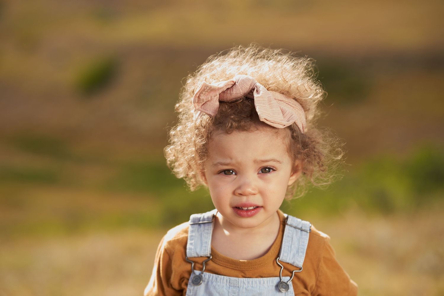 A toddler in overalls and a pink bow smiles at the camera in an outdoor setting.
