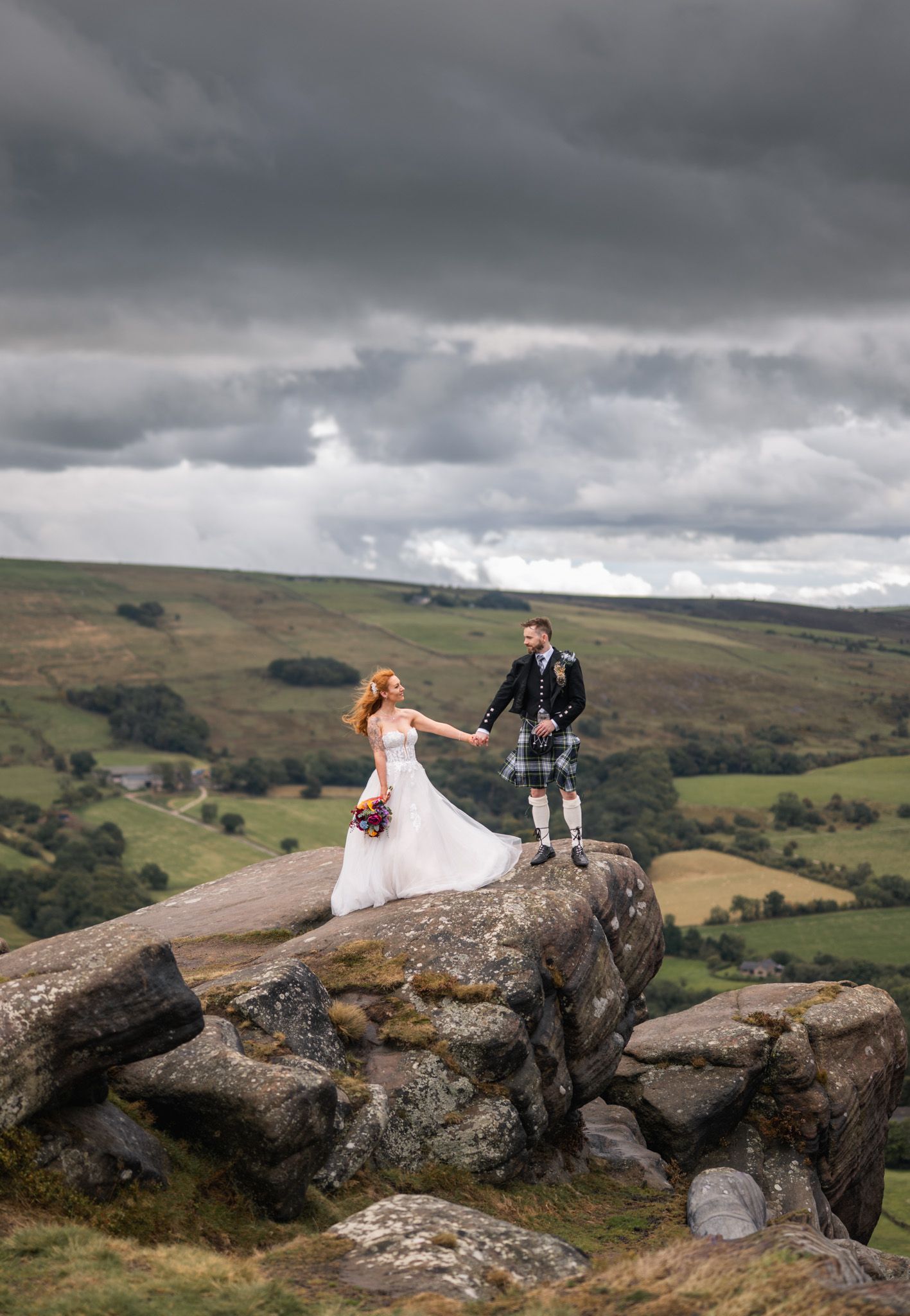 Bride and kilted groom stand atop rocky moorland under dramatic storm clouds in a scenic landscape.