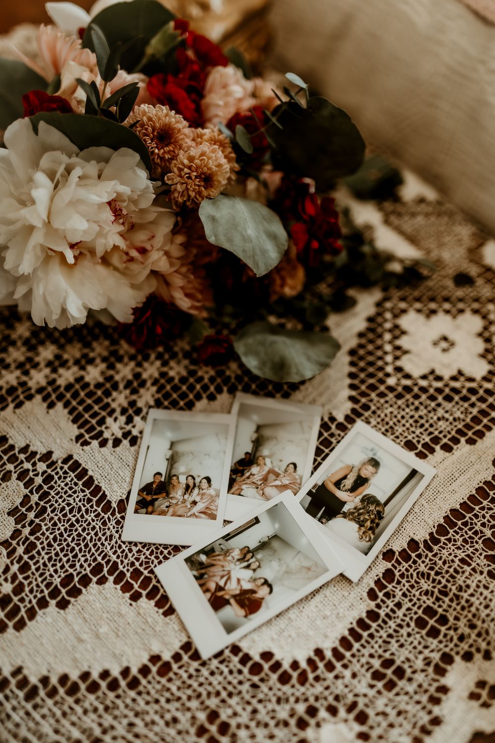 Polaroid photos and a floral bouquet with peonies and greenery arranged on a vintage lace tablecloth.