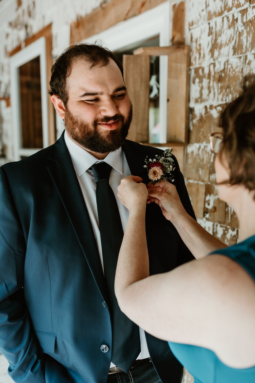 A bearded groom in a dark suit smiles as a woman pins a floral boutonniere to his lapel.