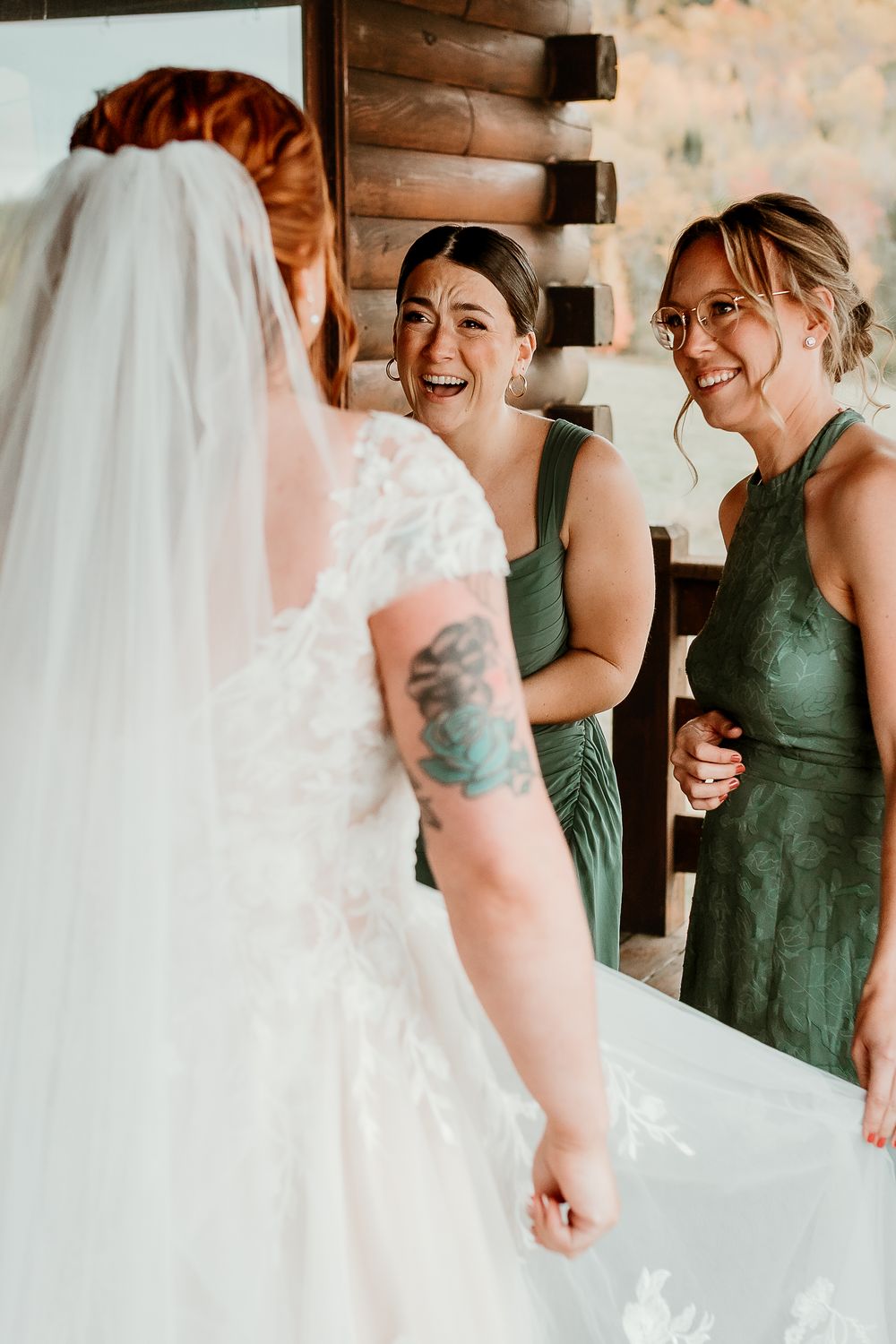 Bride in lace wedding gown and veil shares a joyful moment with two bridesmaids in green dresses.