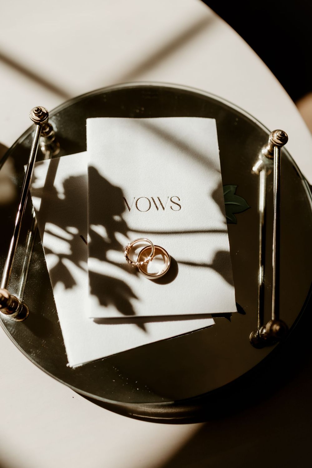 Gold wedding rings resting on white vows booklets on a decorative tray with elegant shadows.