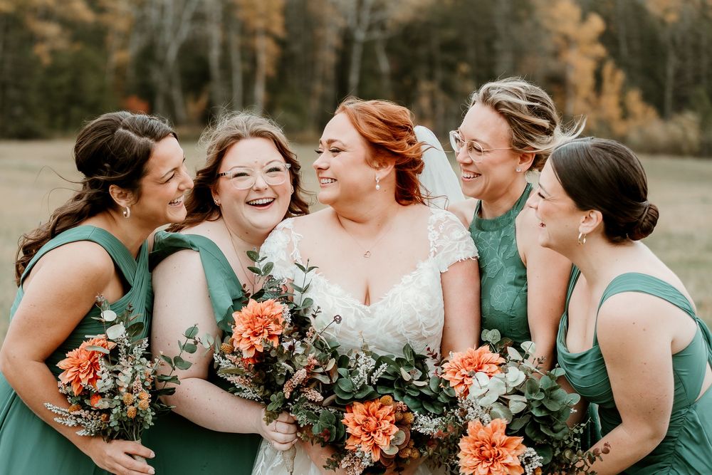 Bride in white lace gown laughs with bridesmaids in green dresses holding orange floral bouquets outdoors.