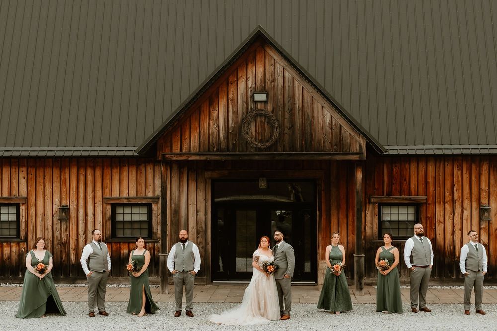Wedding party poses before rustic wooden barn, bridesmaids in sage green gowns, groomsmen in gray suits, bride in white dress.