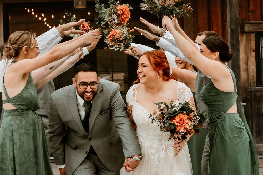 Bride and groom walk through floral arch tunnel with wedding party in sage green dresses at rustic barn venue.