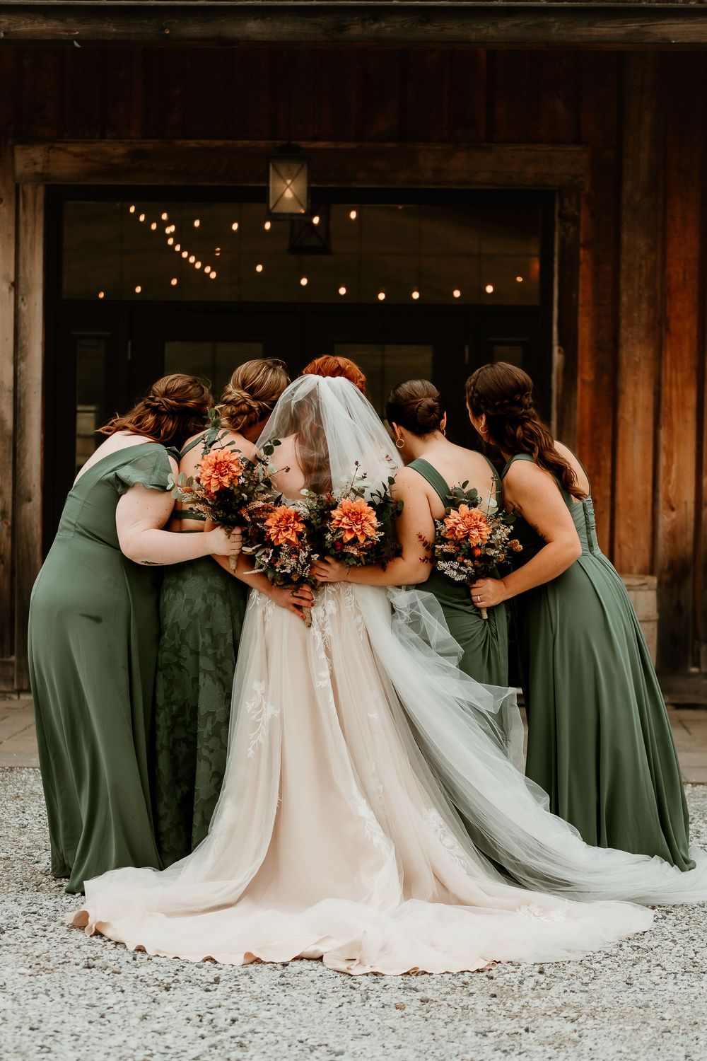 Bride and bridesmaids in sage green gowns holding orange bouquets, posing from behind outside a rustic barn venue.