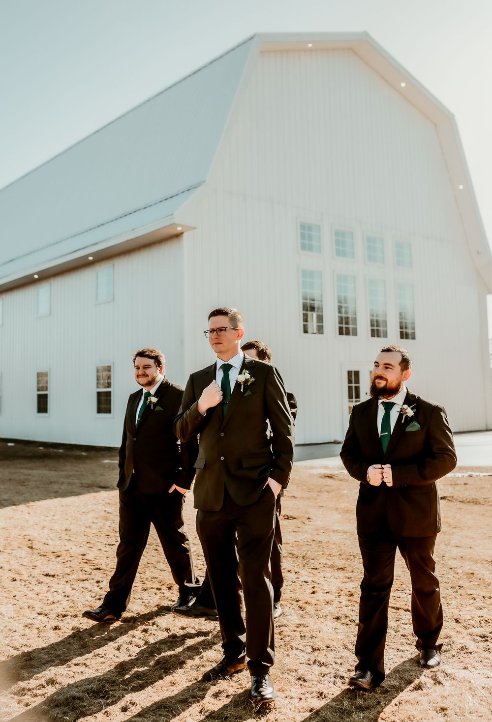 Groom and groomsmen in dark suits with green ties walk outside a white barn wedding venue.
