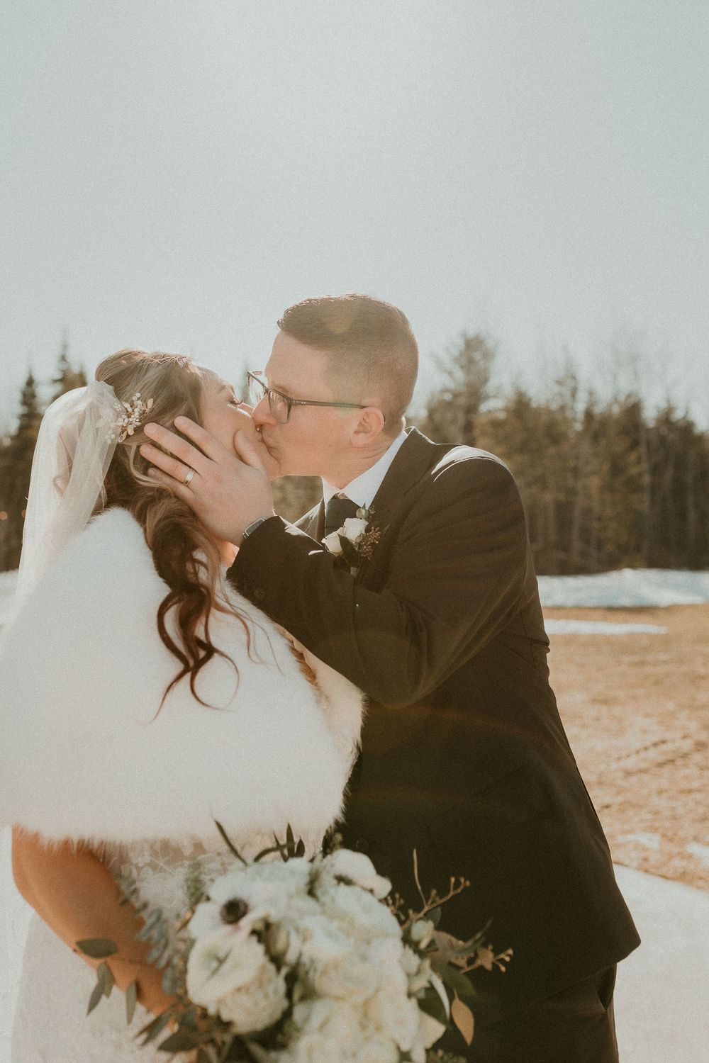 A bride and groom share a romantic kiss outdoors in winter, with the bride holding a white bouquet and wearing a fur wrap.