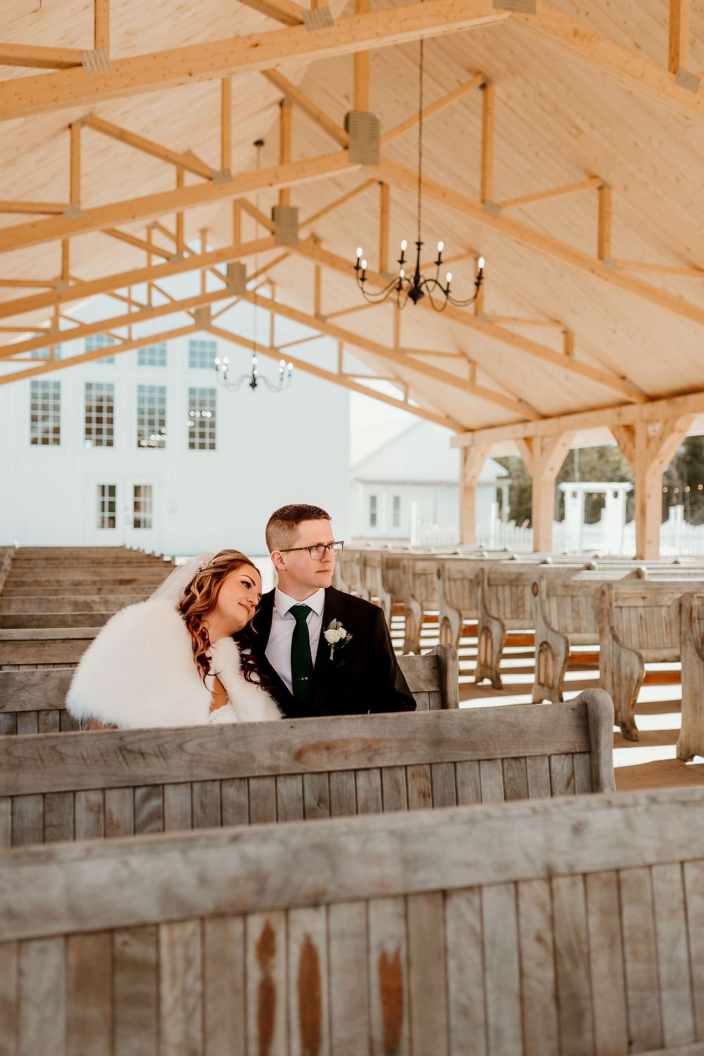 Bride and groom sitting together on rustic wooden pews under an open-air chapel with exposed timber beams.
